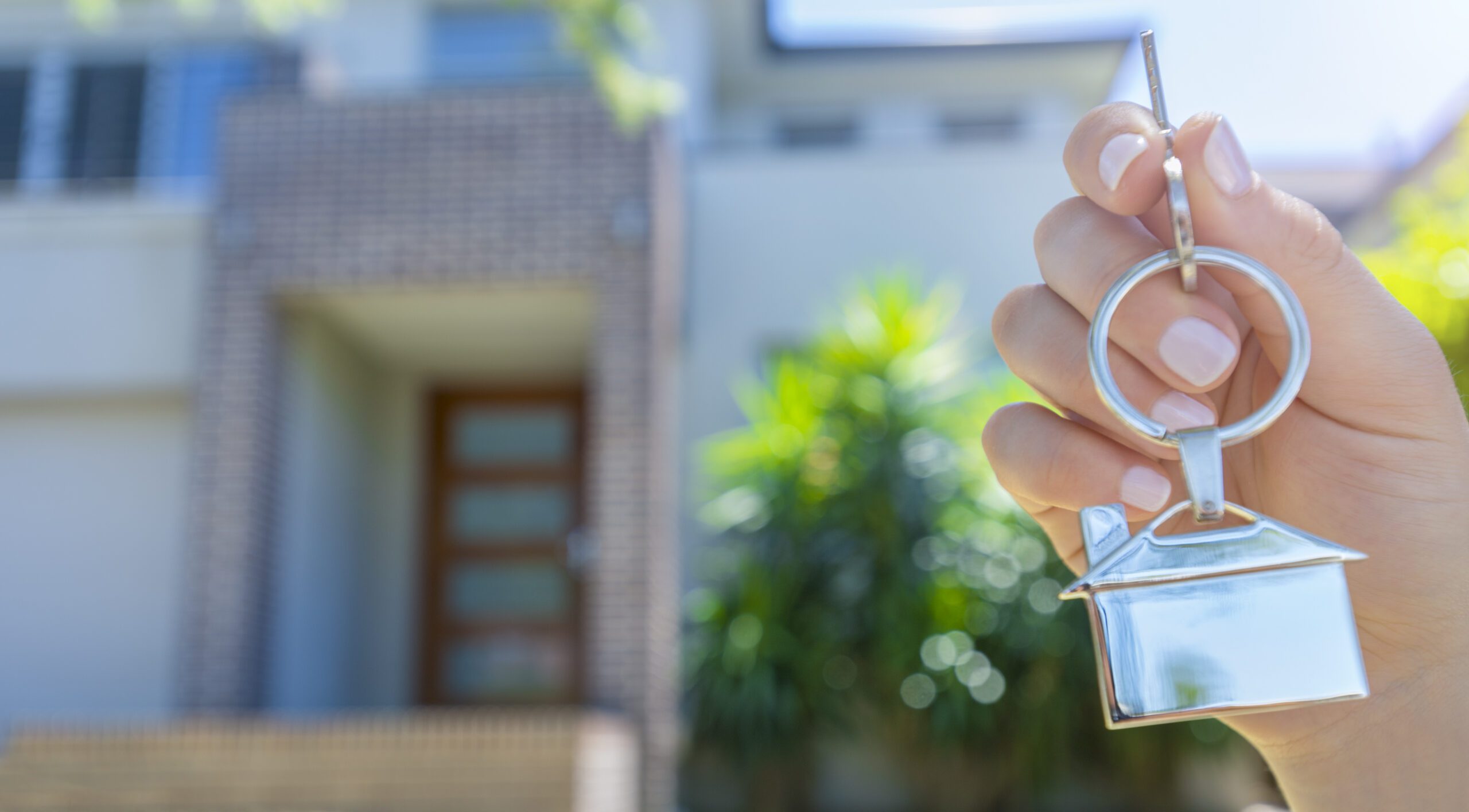 Hand holding a house key in front of a large house.