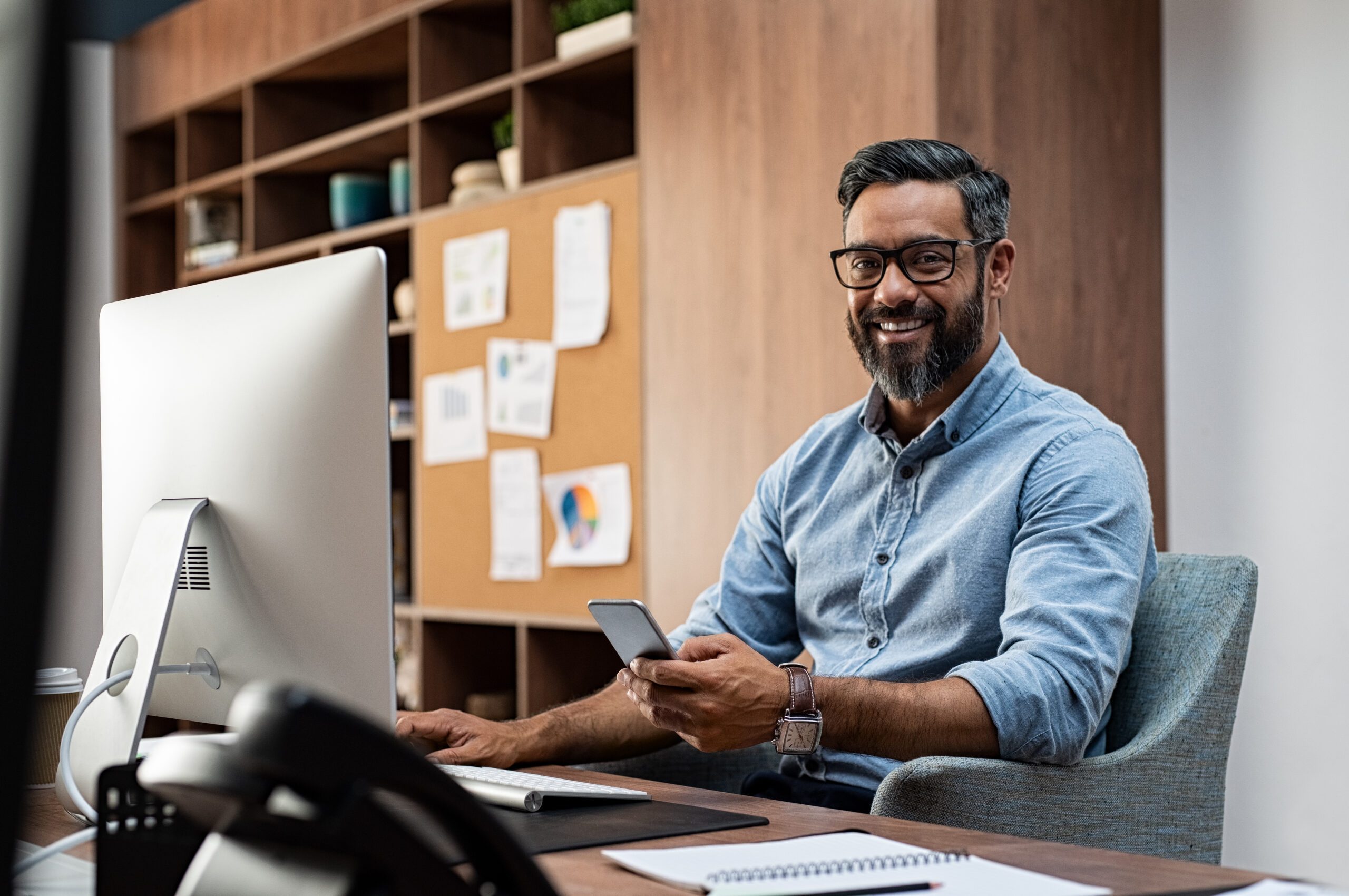 Smiling business man wearing eyeglasses working on desktop computer while using phone in office